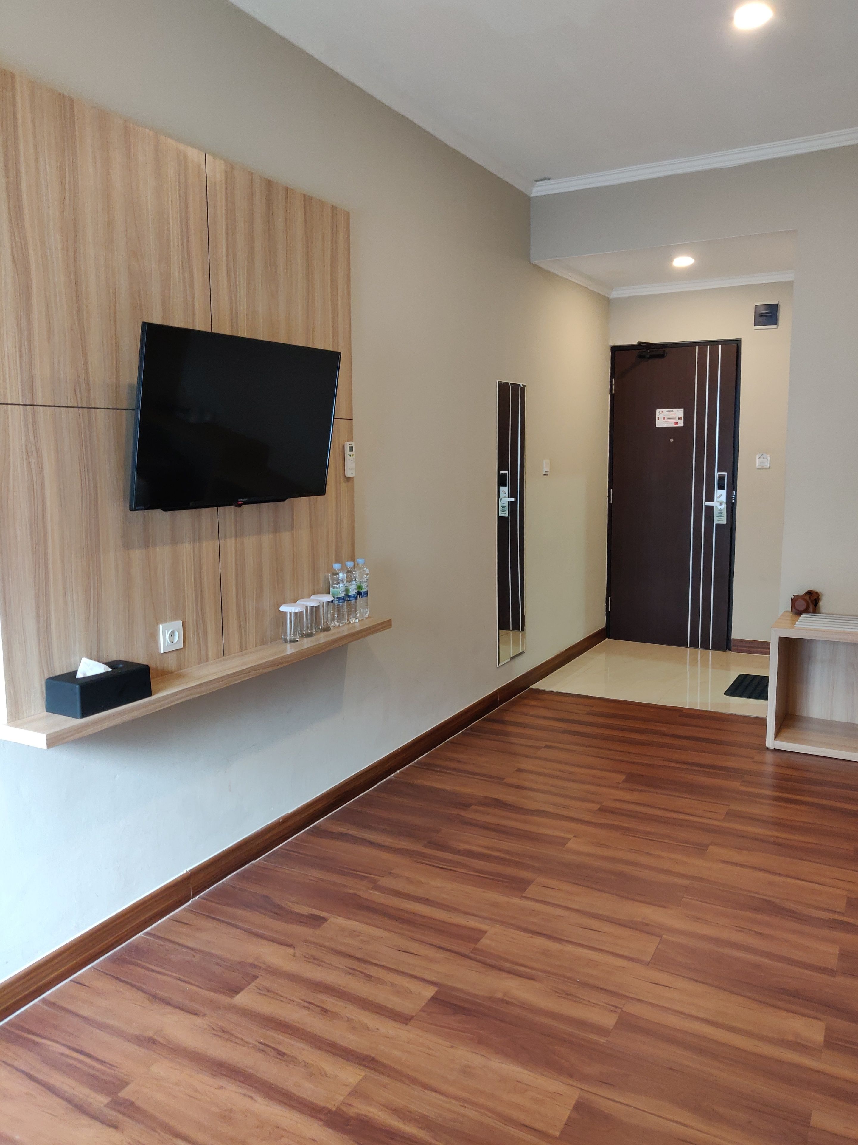A spacious room with a wooden floor, a mounted flat-screen TV, a shelf with water bottles, and a door with a sign.
