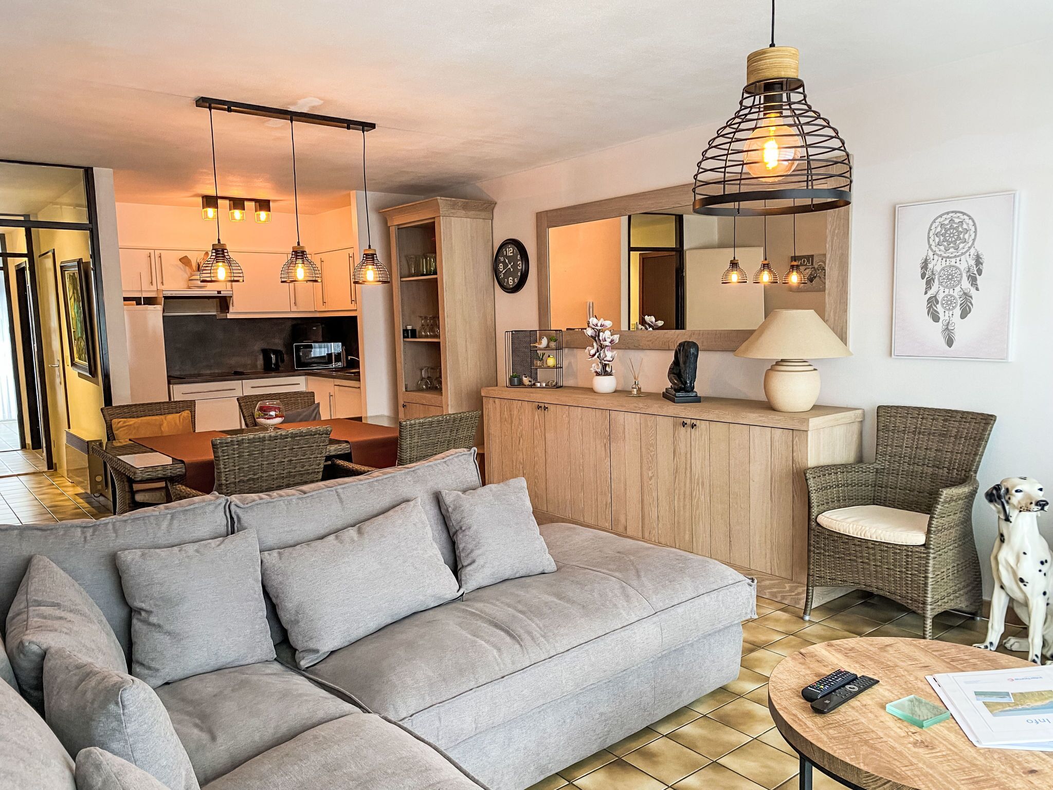 Ett modernt vardagsrum med en grå soffa, flätade stolar, en matplats och ett kök i bakgrunden.