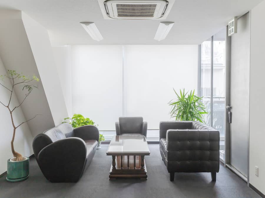 A modern office space with a sofa, armchairs, and a coffee table. There are potted plants and a large window providing natural light.
