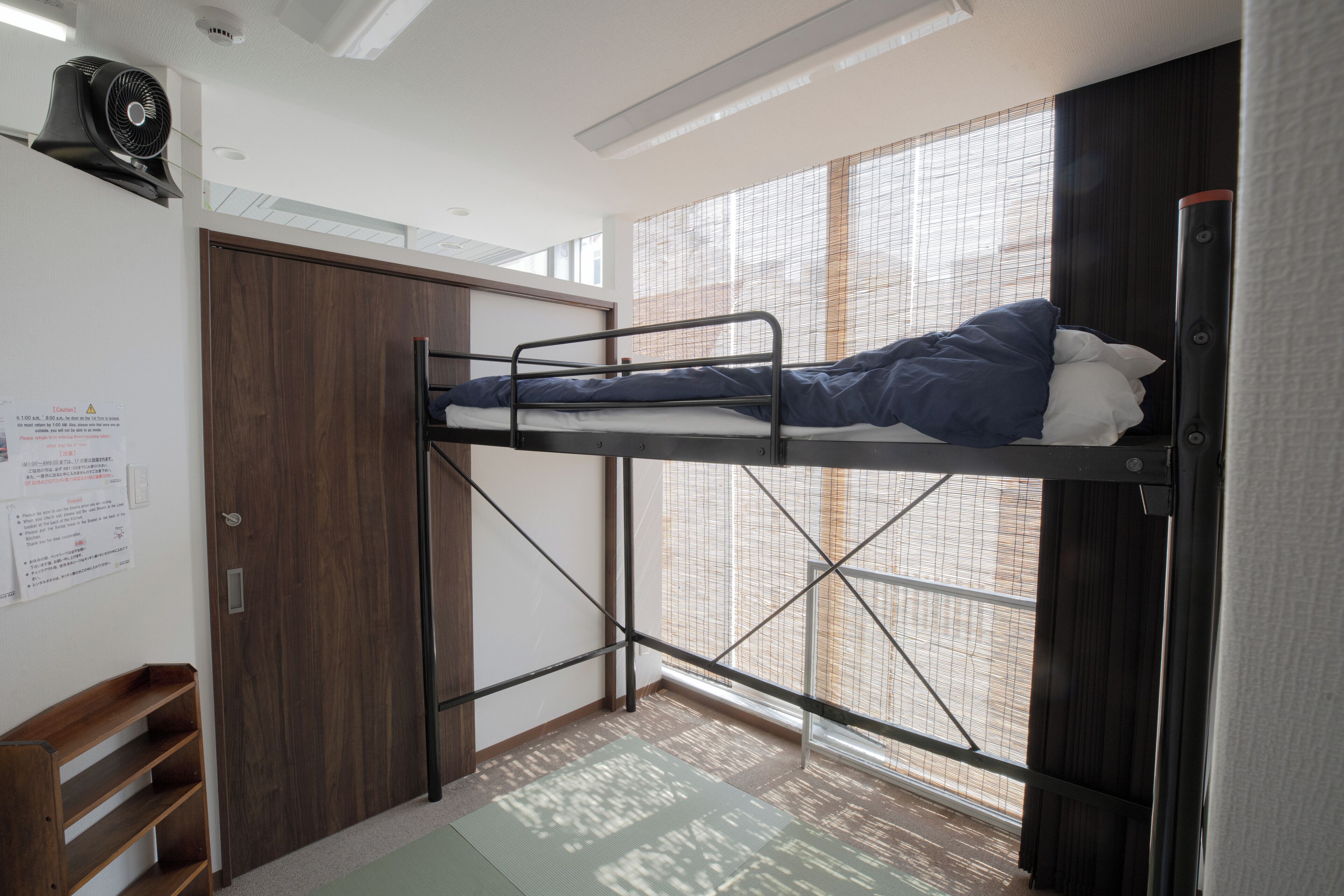 A bunk bed room with a wooden wardrobe, a fan, and a window with blinds.