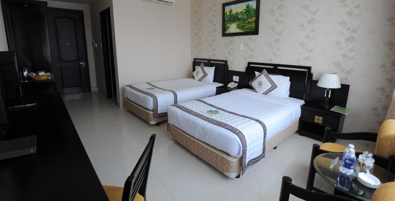 A hotel room with two single beds, a desk, a chair, a TV, and a small table with a lamp and a bottle.