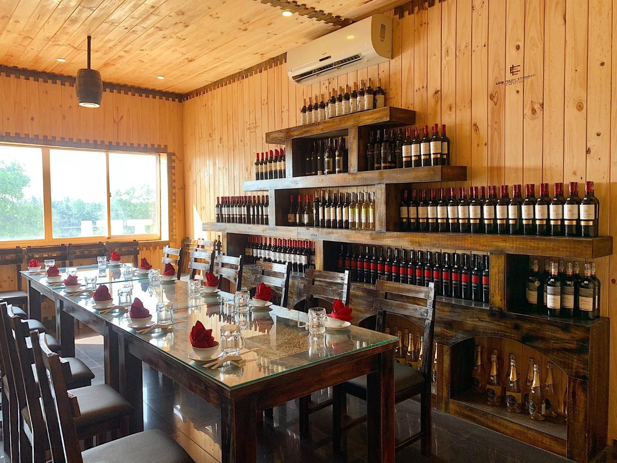 A restaurant interior with a long dining table set for multiple guests, wooden walls, and a wine rack filled with bottles.