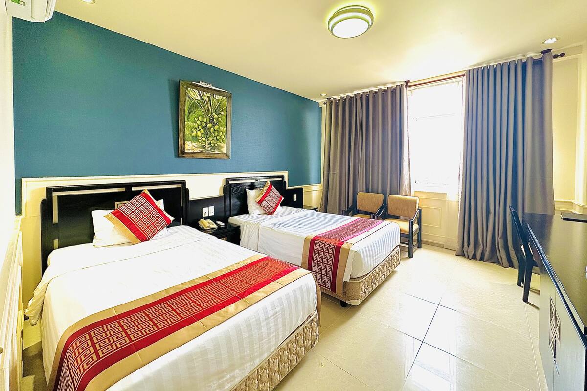 Habitación de hotel con dos camas, una mesita, una silla, ventana con cortinas e iluminación en el techo.