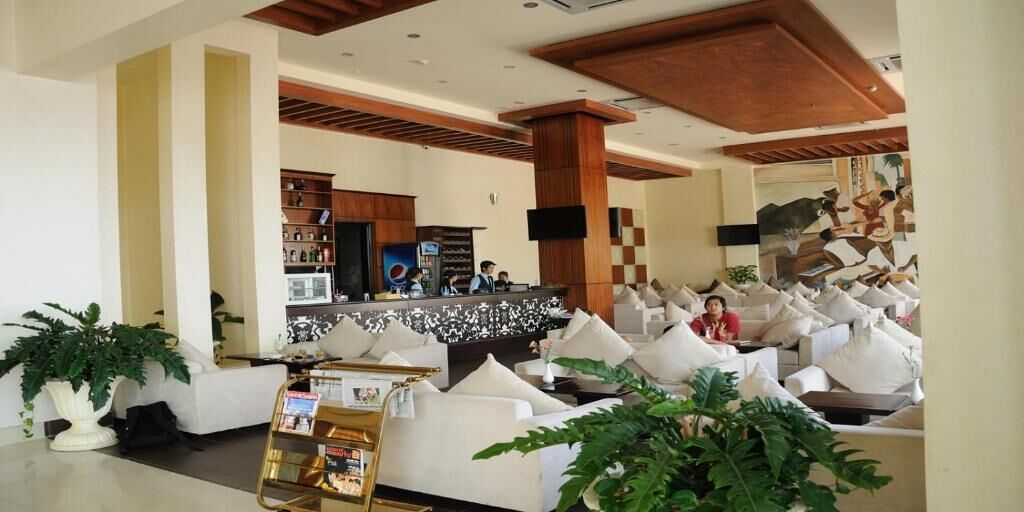 Un hall d’hôtel comprenant un comptoir d’accueil, un espace détente avec des canapés et des coussins blancs, ainsi que des plantes en pot.