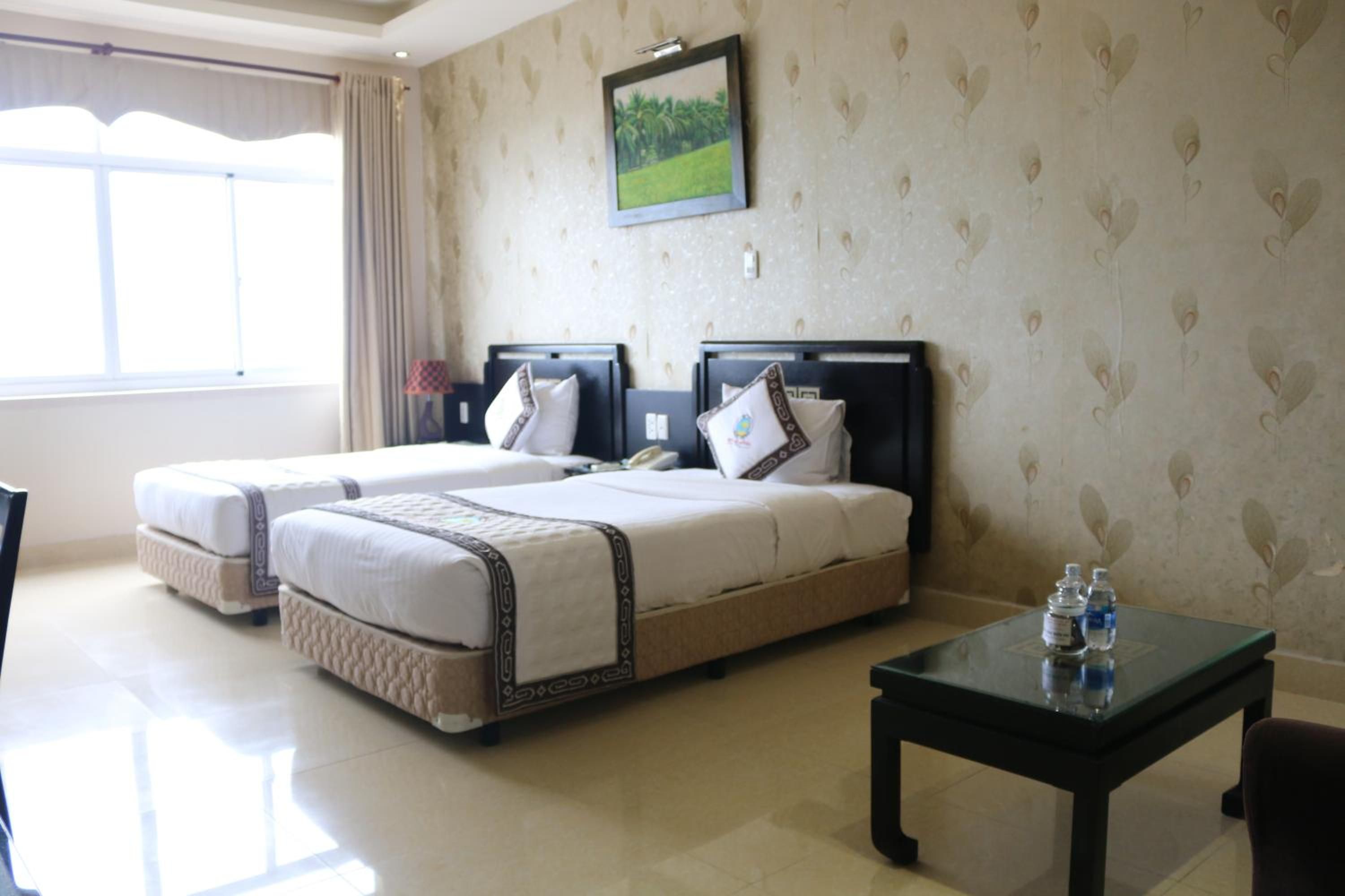 A hotel room with two beds, a nightstand, a table with water bottles, and a window with curtains.