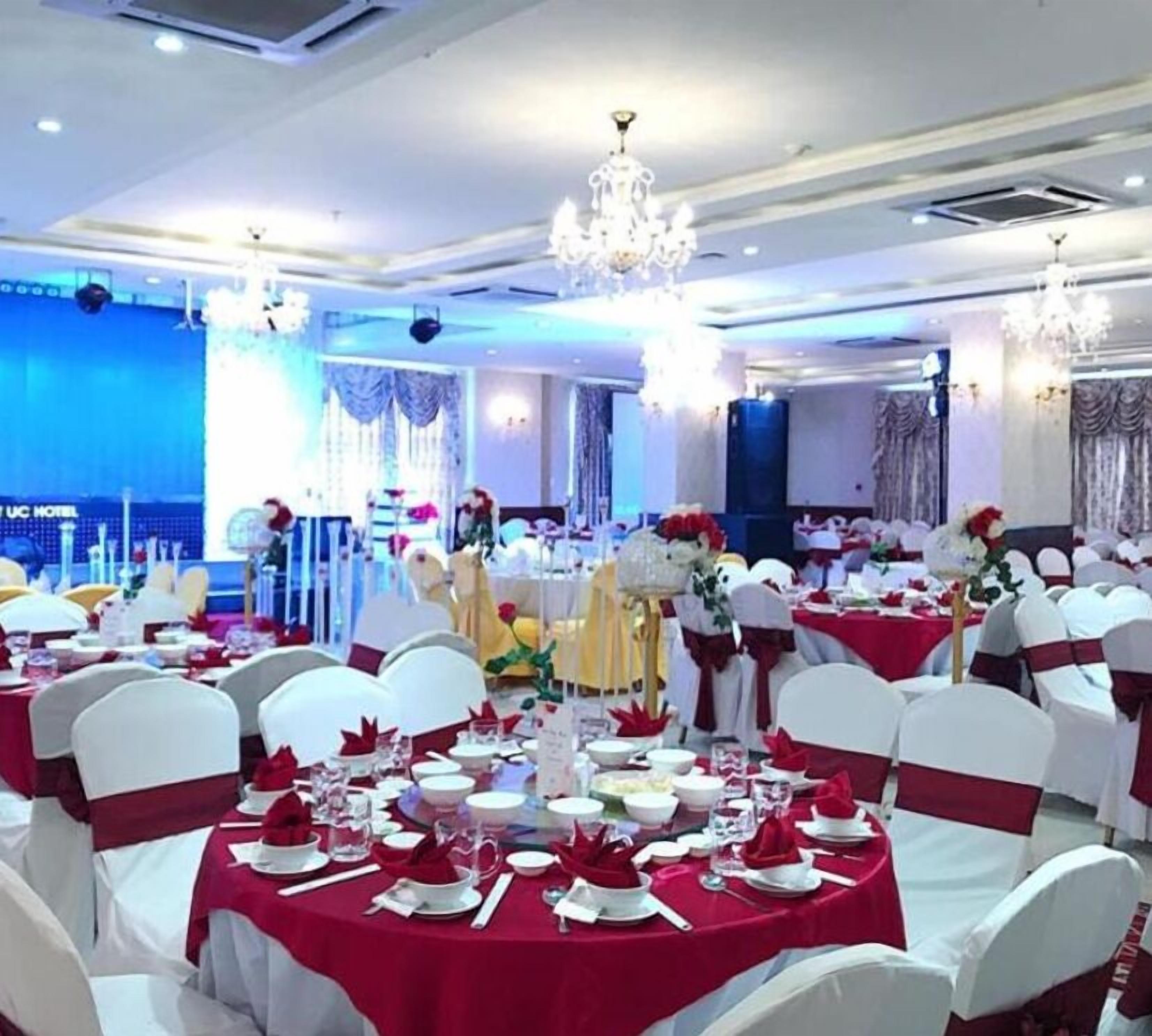 A banquet hall with round tables covered in red tablecloths, white chairs with red sashes, and chandeliers.