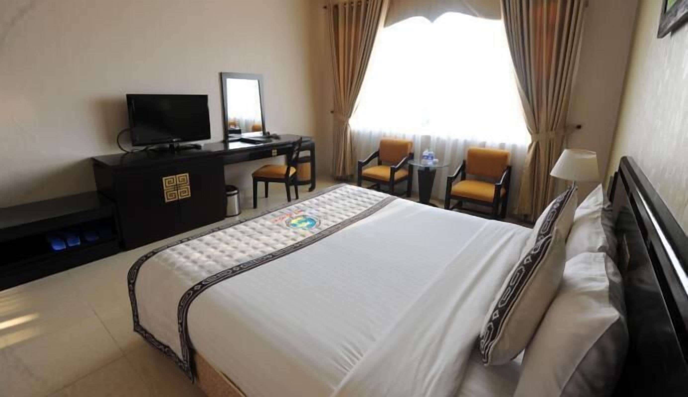 A hotel room with a bed, a desk with a TV, two chairs, a small table, and a window with curtains.
