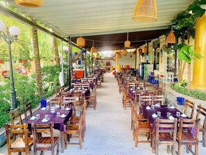 Un restaurante al aire libre con filas de mesas y sillas, rodeado de follaje y un paseo techado.