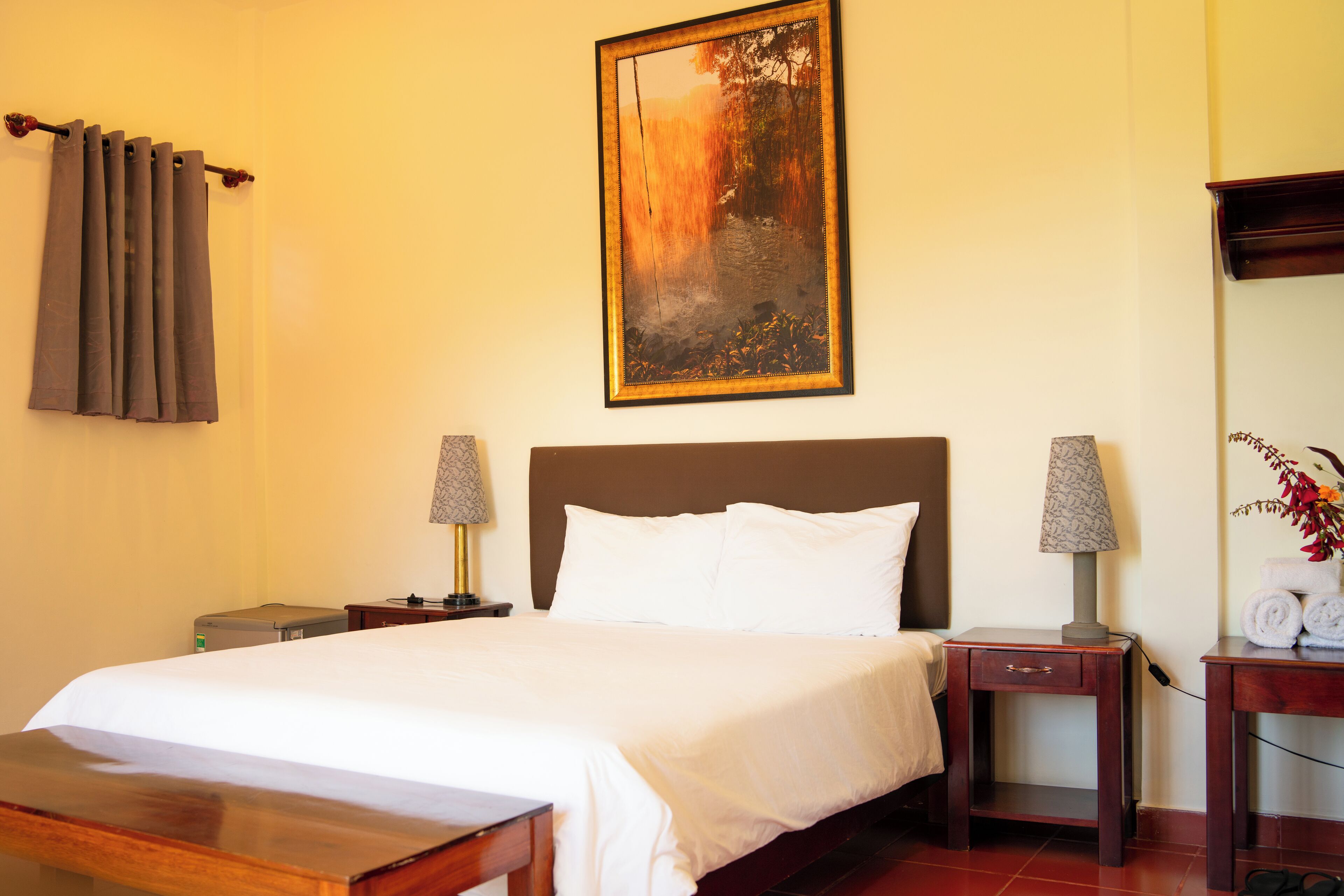 A hotel room with a large bed, two bedside tables with lamps, a small table with towels, and a painting on the wall.