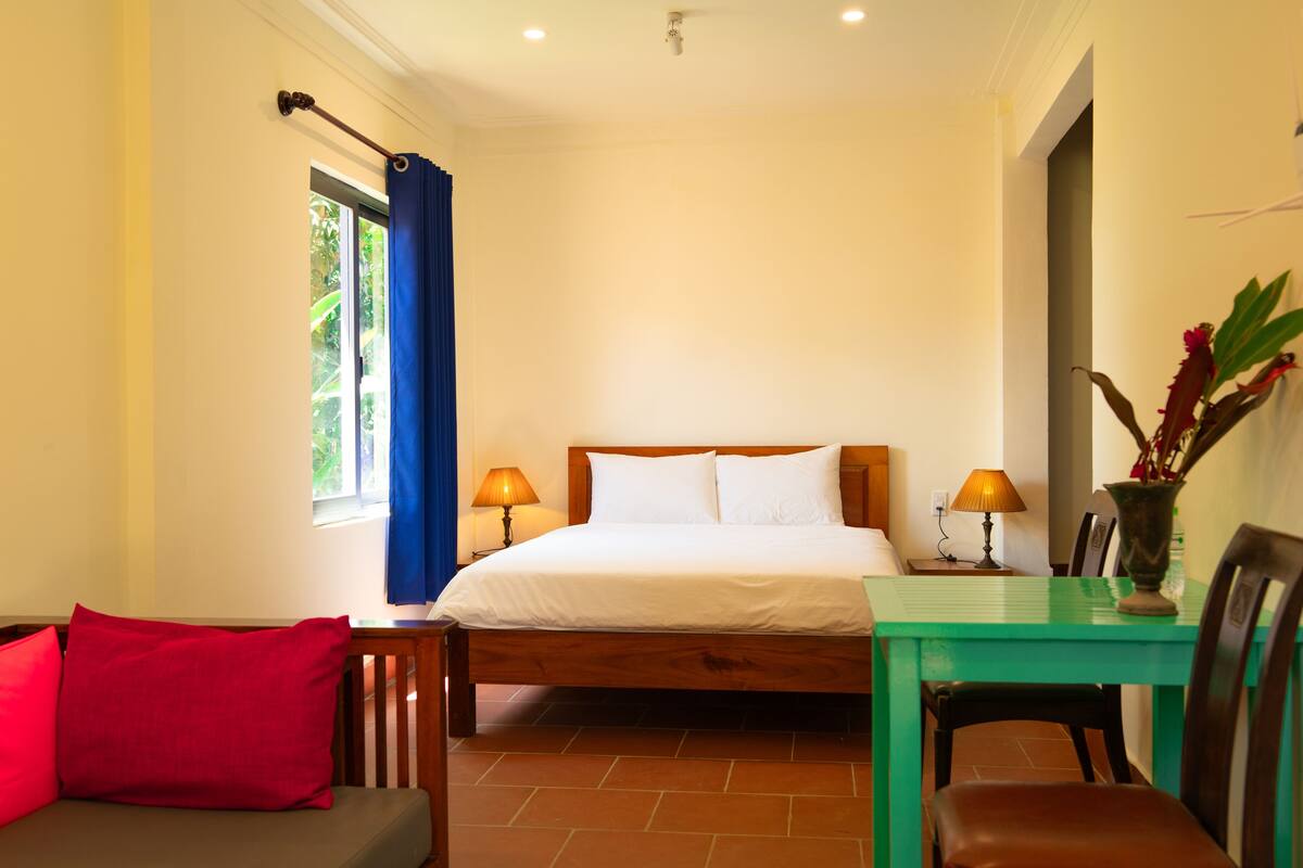 A bedroom with a bed, a table, a chair, a window with curtains, and a plant.
