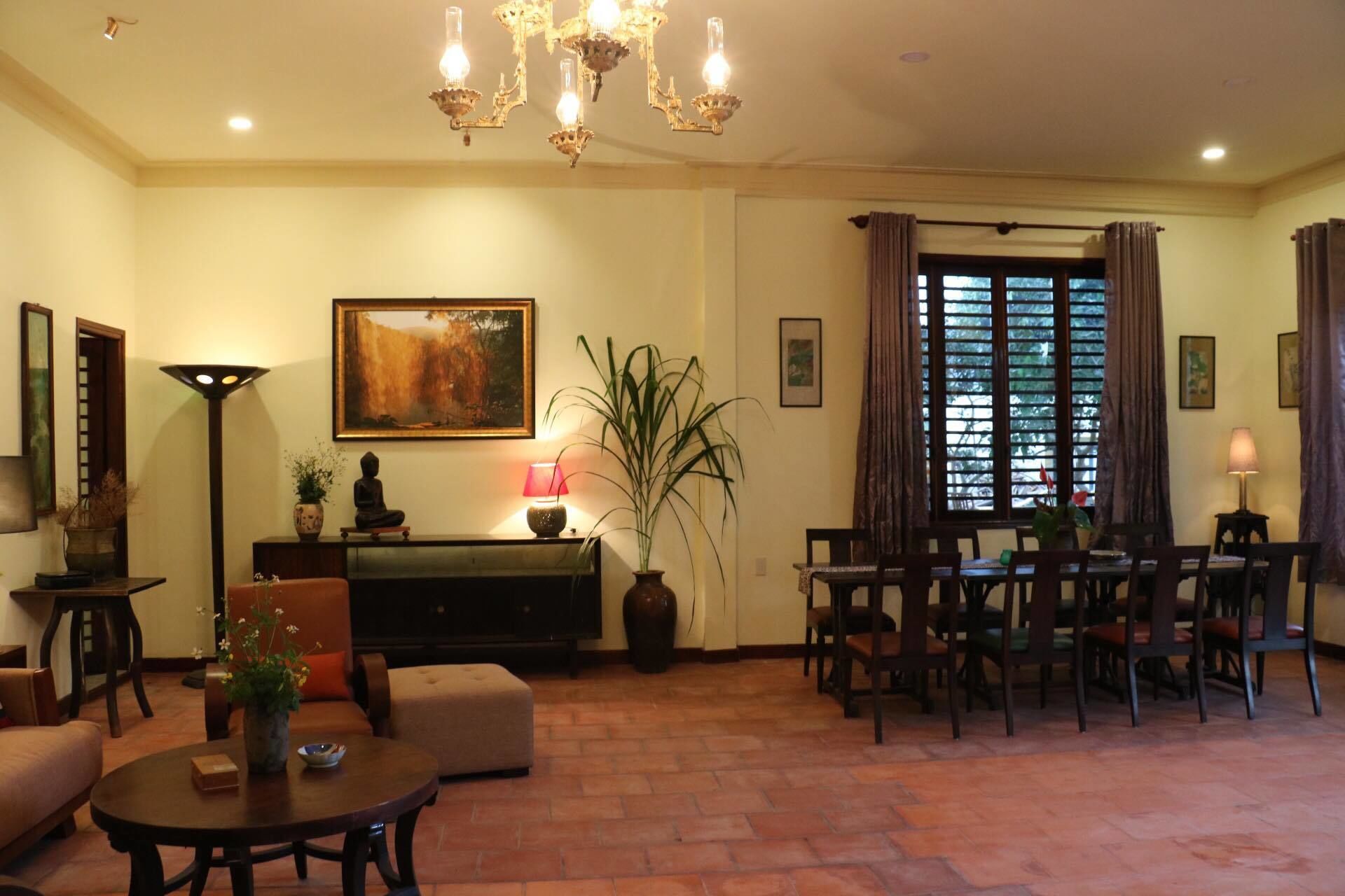 Una sala de estar con un sofá, una mesa de centro, una mesa de comedor con sillas, una araña, un cuadro y una ventana con cortinas.