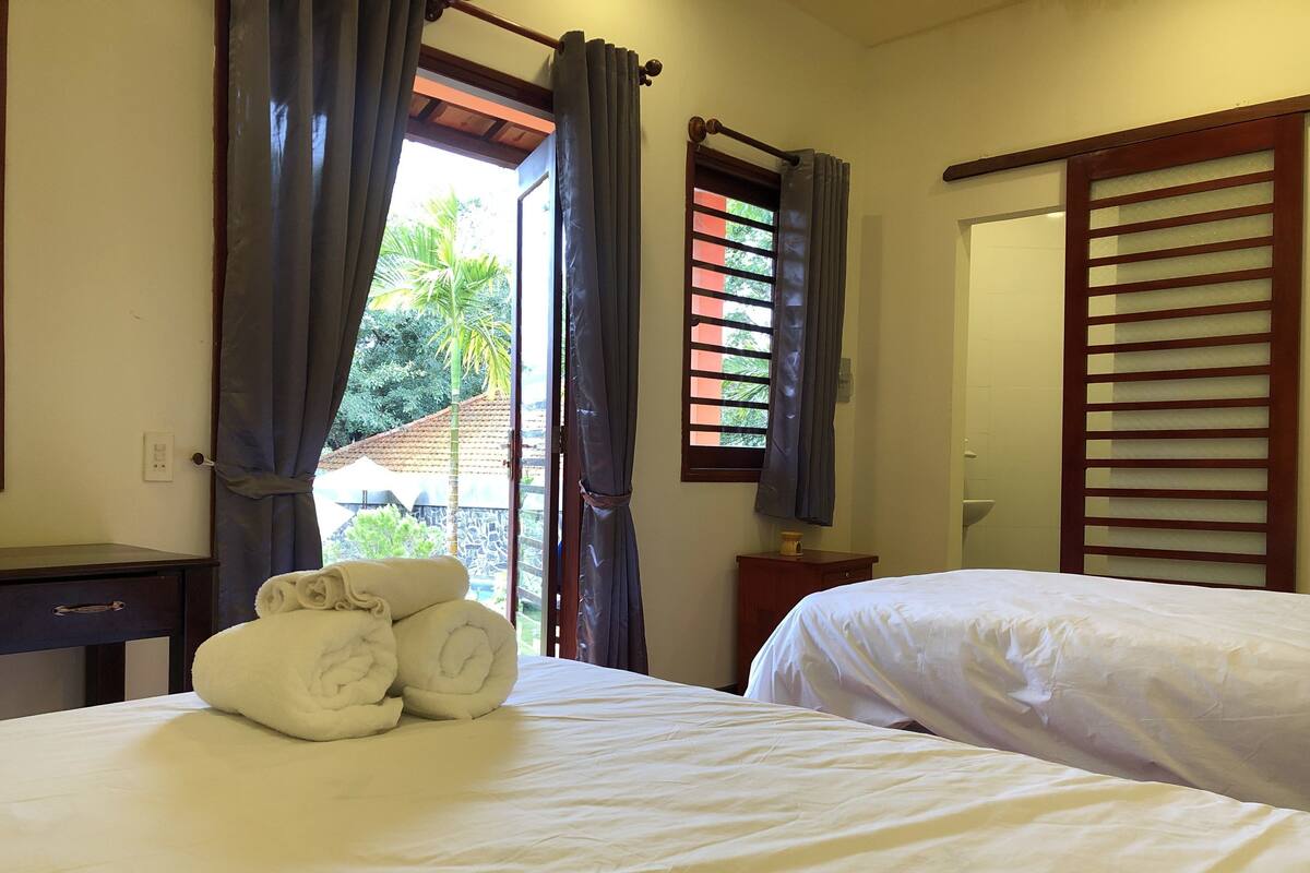 A hotel room with two beds, a small table, a mirror, and a window with curtains.