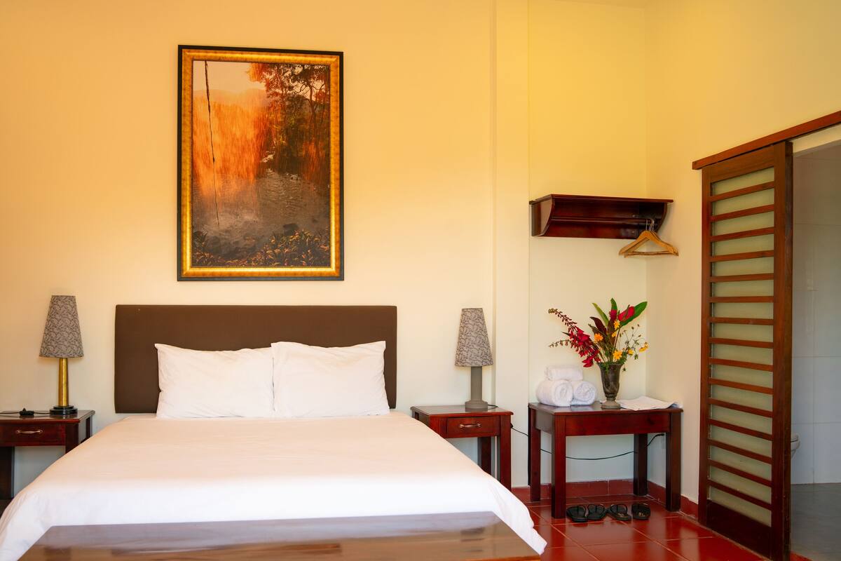 A neatly made bed with white linens, a framed painting above it, two bedside tables with lamps, a small table with a vase of flowers, and a wooden wardrobe.