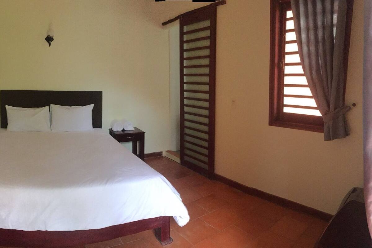 A bedroom with a bed, a nightstand, a wall-mounted shelf, a window with blinds, and a door with a shutter.