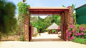 Una entrada techada con estructura de techo de paja, pilares de piedra y un sendero que lleva a un edificio.
