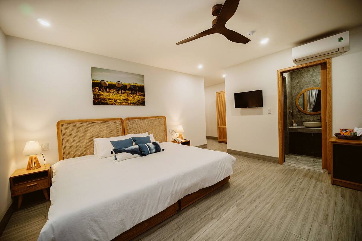 A hotel room with a large bed, a ceiling fan, a mounted TV, and a bathroom with a mirror and sink.