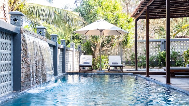 A swimming pool with a waterfall feature, two lounge chairs, and a pergola with an umbrella. - The Watermark Beachside Hotel (Da Nang)