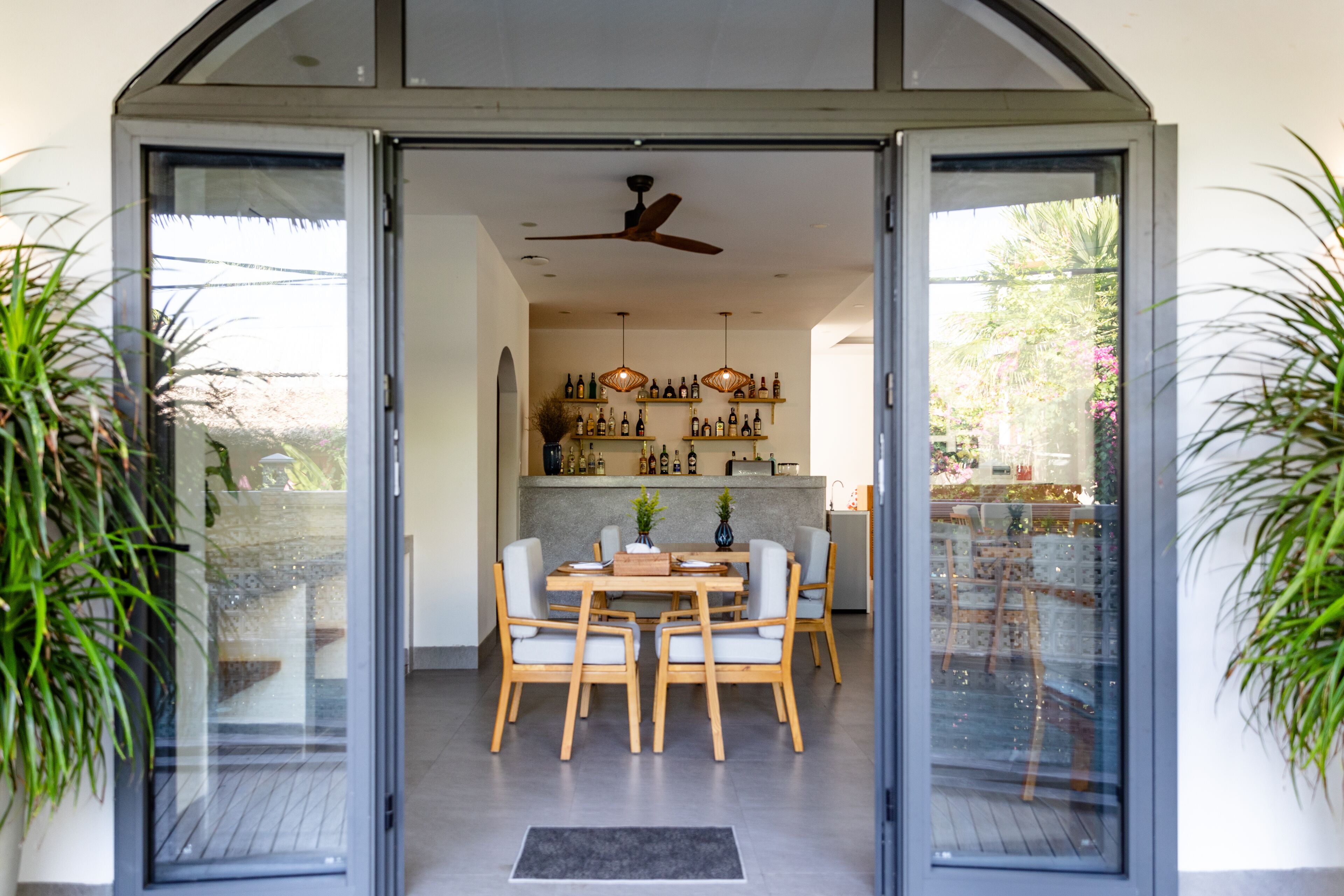 A modern dining area with a glass door entrance, a ceiling fan, and a kitchen with shelves of bottles in the background.