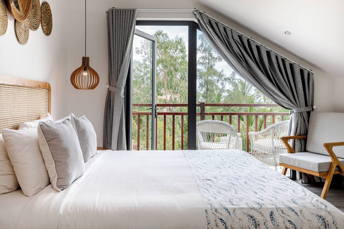 A bedroom with a large bed, a balcony with wicker furniture, and a view of trees.
