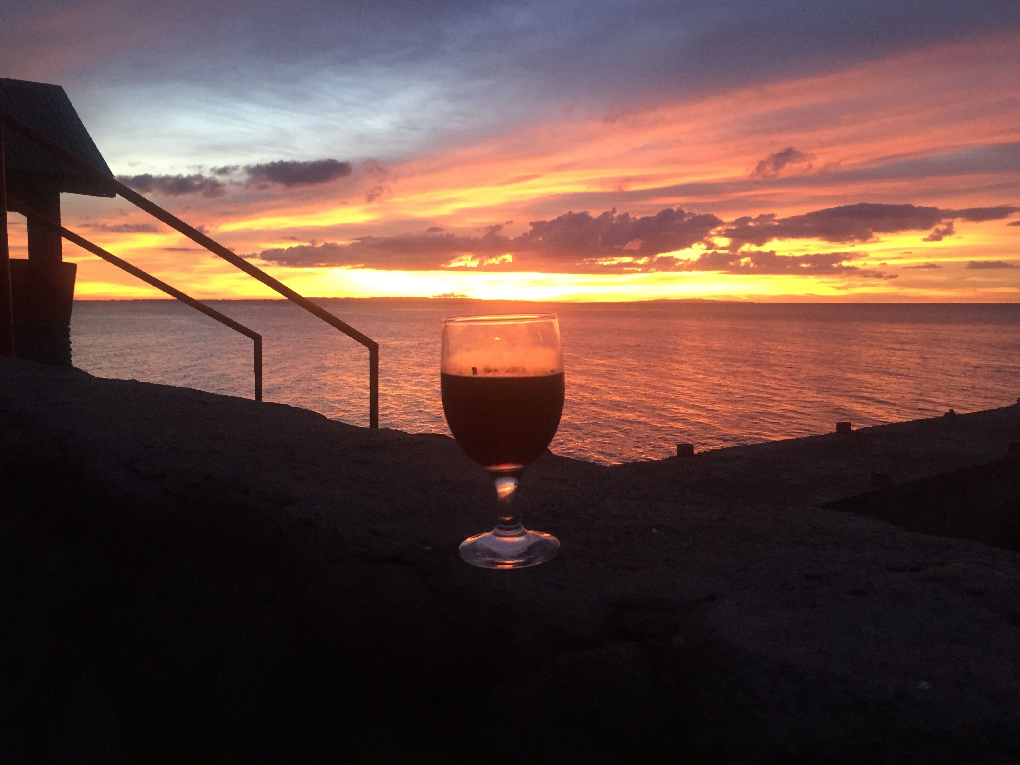A glass of red wine on a balcony overlooking a sunset over the ocean.