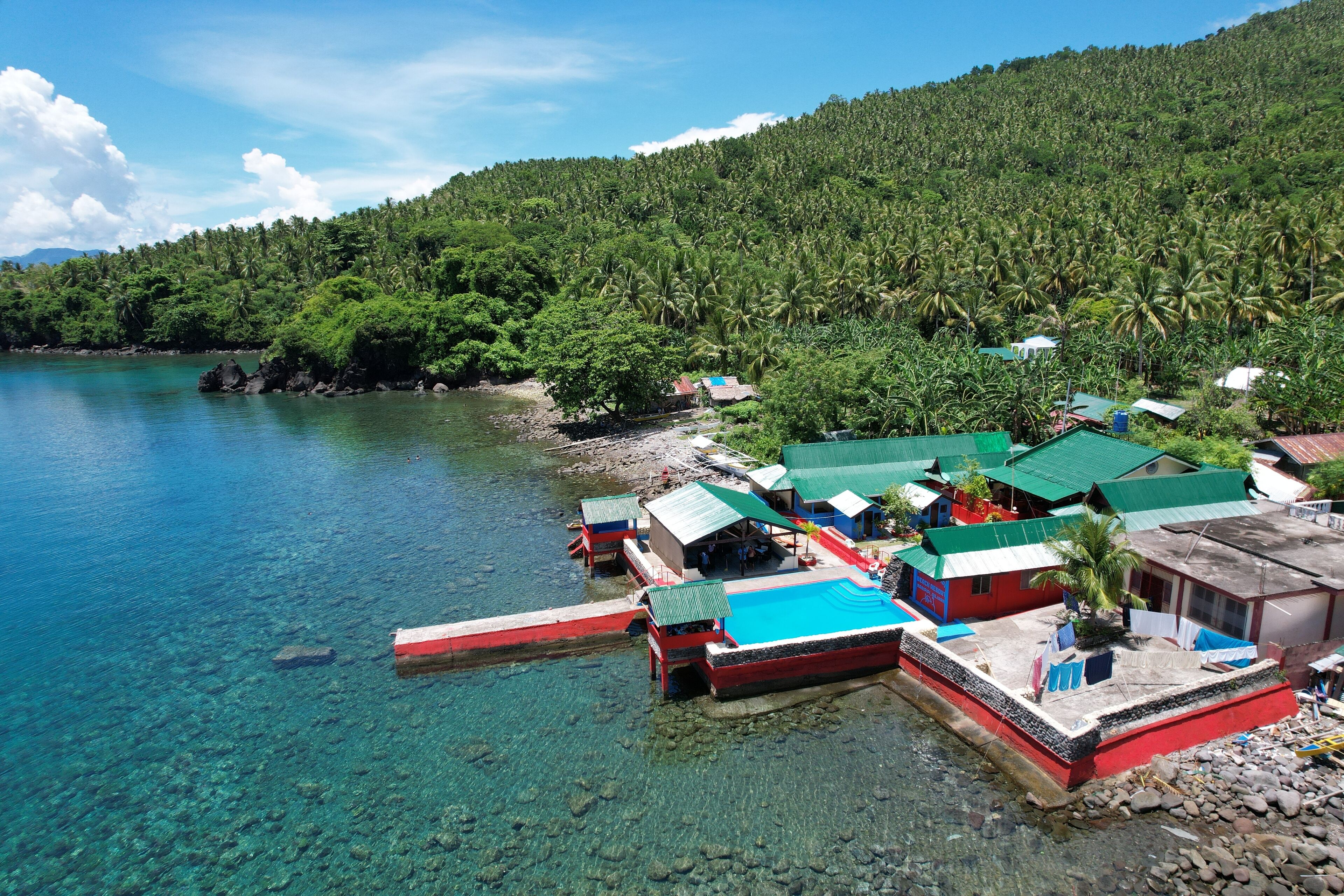 A coastal area with clear waters, a dock with several small buildings, and a lush green hillside in the background.