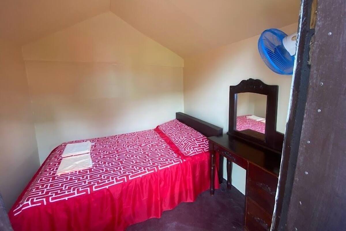 Ein kleines Zimmer mit einem Einzelbett, einer roten Bettdecke, einem Nachttisch aus Holz, einem Spiegel und einem blauen Ventilator.