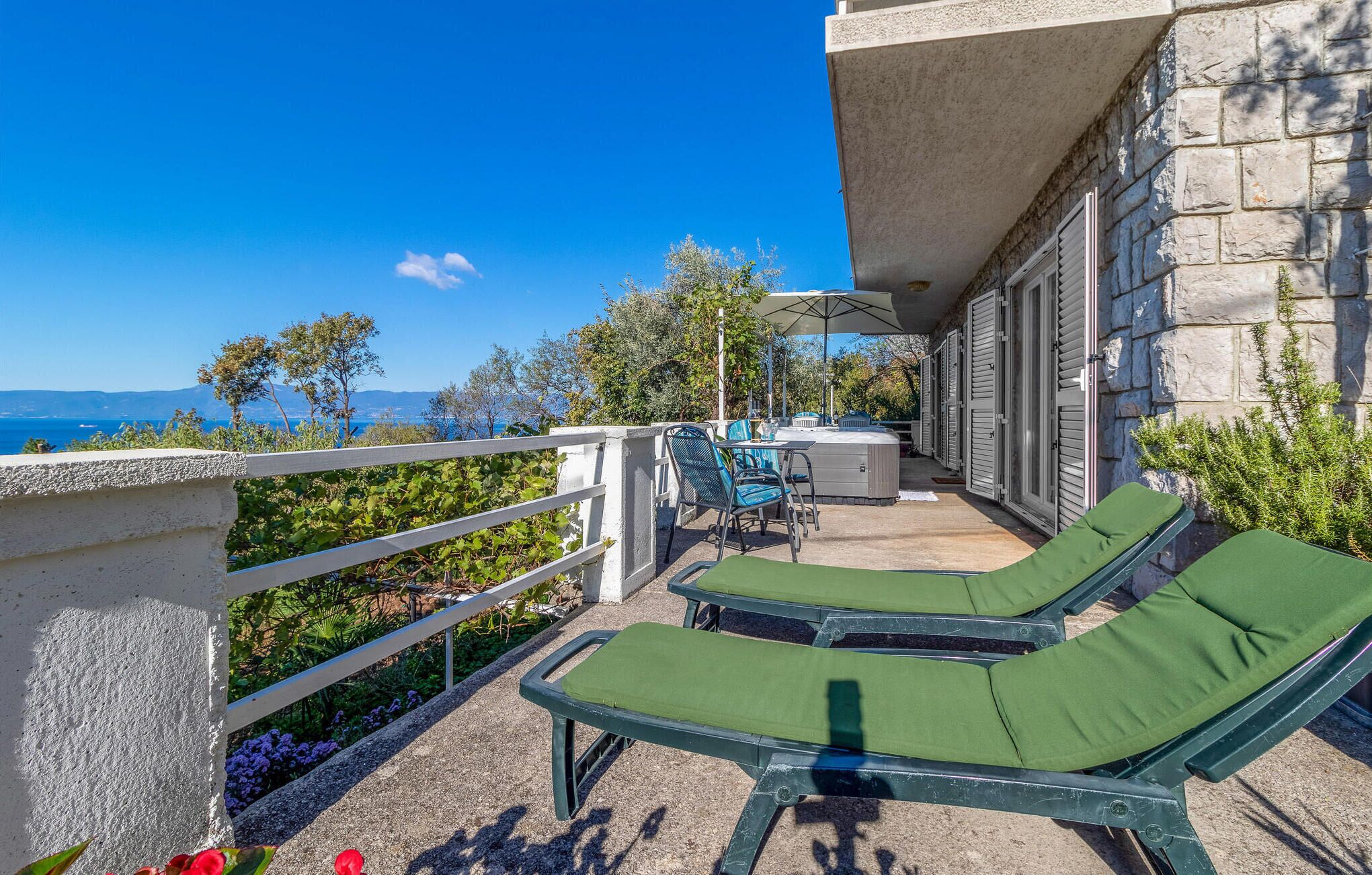 A balcony with two green lounge chairs, a table with chairs, and a view of the sea and mountains.