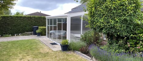 A conservatory with a glass roof, surrounded by a well-maintained garden with various plants and a hedge.