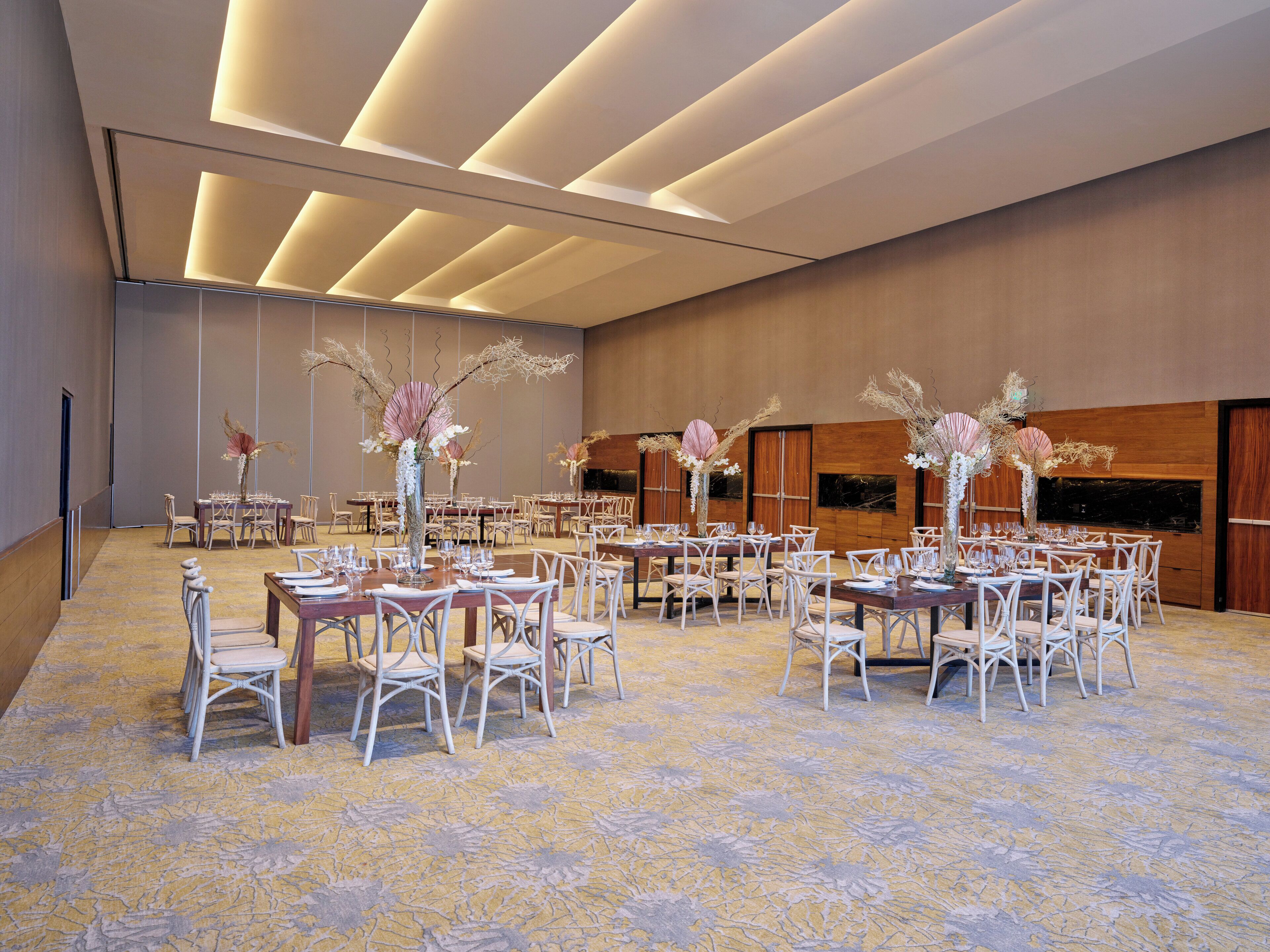 A spacious banquet hall with multiple tables and chairs, elegant floral centerpieces, and a modern ceiling design.