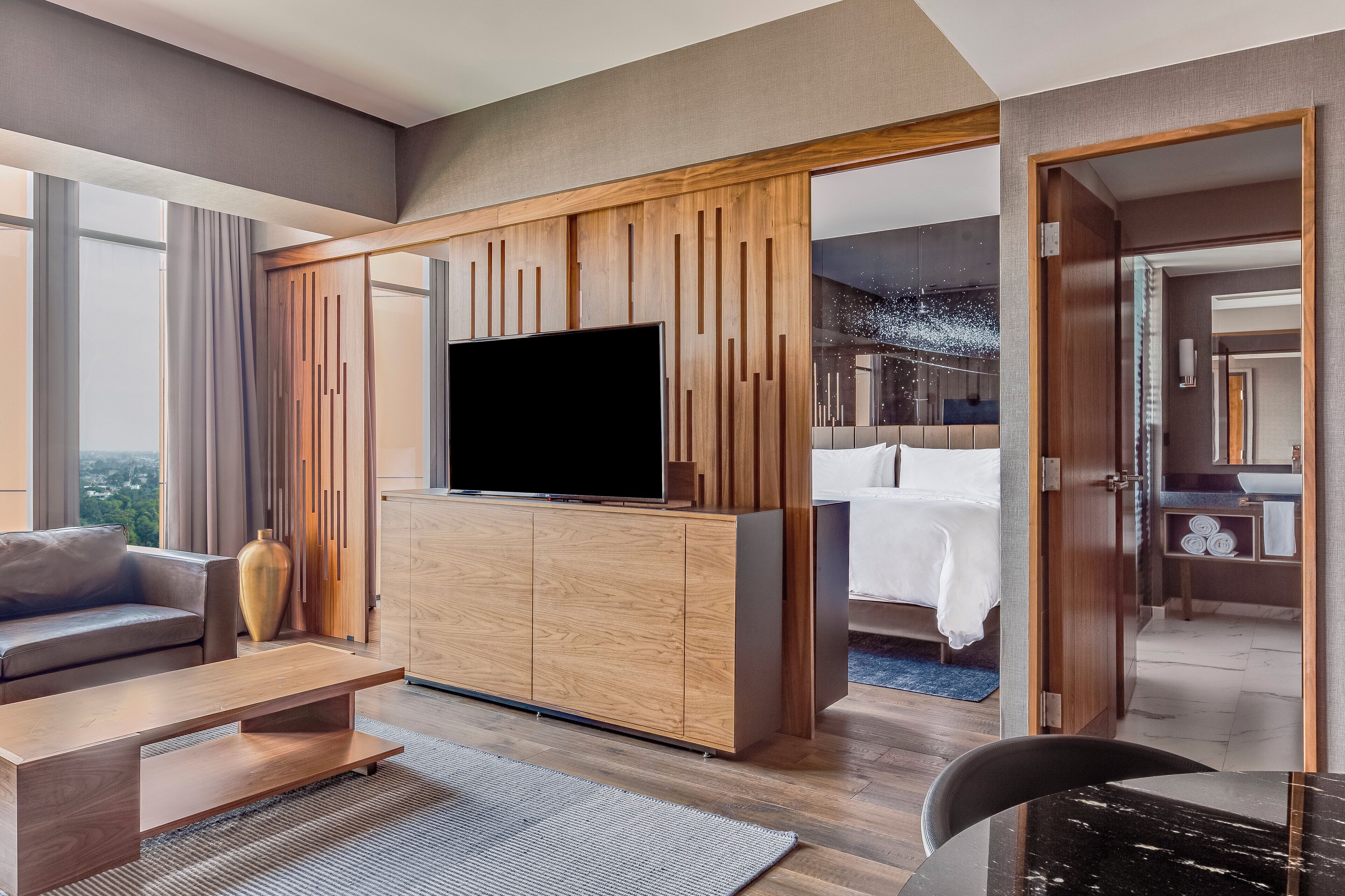 A modern hotel room with a large flat-screen TV, a sofa, a coffee table, and a wooden cabinet.