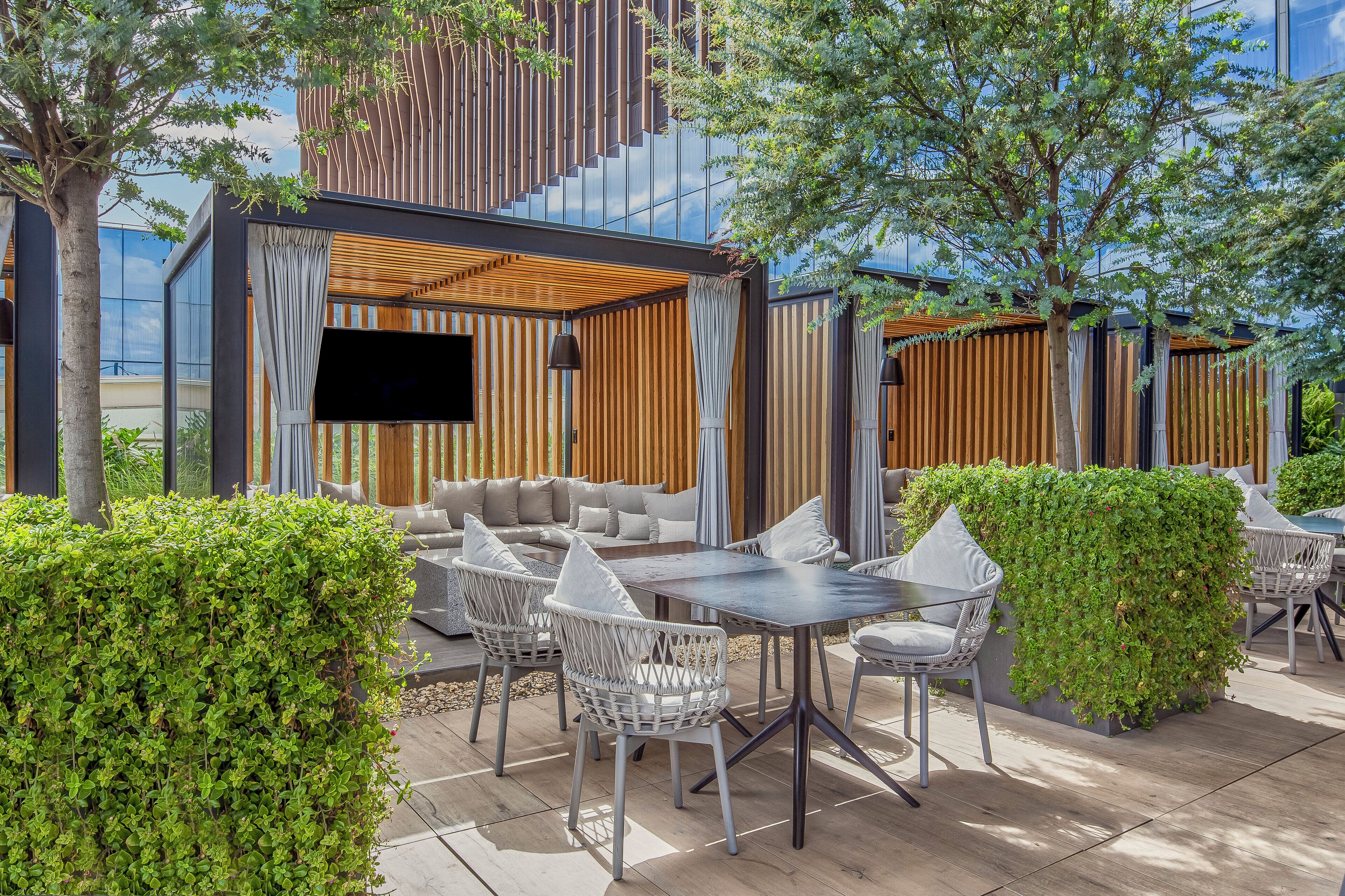 Un espace extérieur aménagé avec un canapé, des fauteuils et une table, entouré de verdure et avec un bâtiment moderne en arrière-plan.