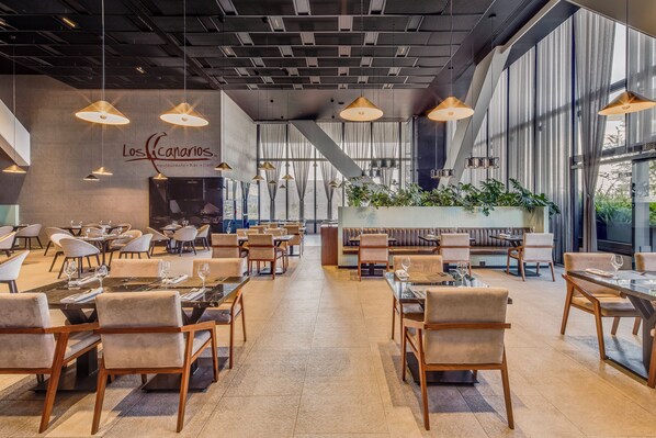 Interior espaçoso de restaurante com várias mesas e cadeiras, iluminação pendente e uma grande janela com cortinas.