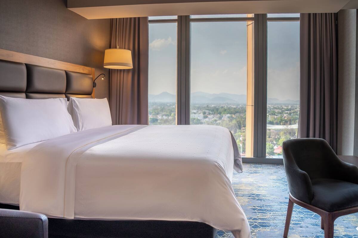 A hotel room with a large bed, a chair, a lamp, and a view of a cityscape through large windows.