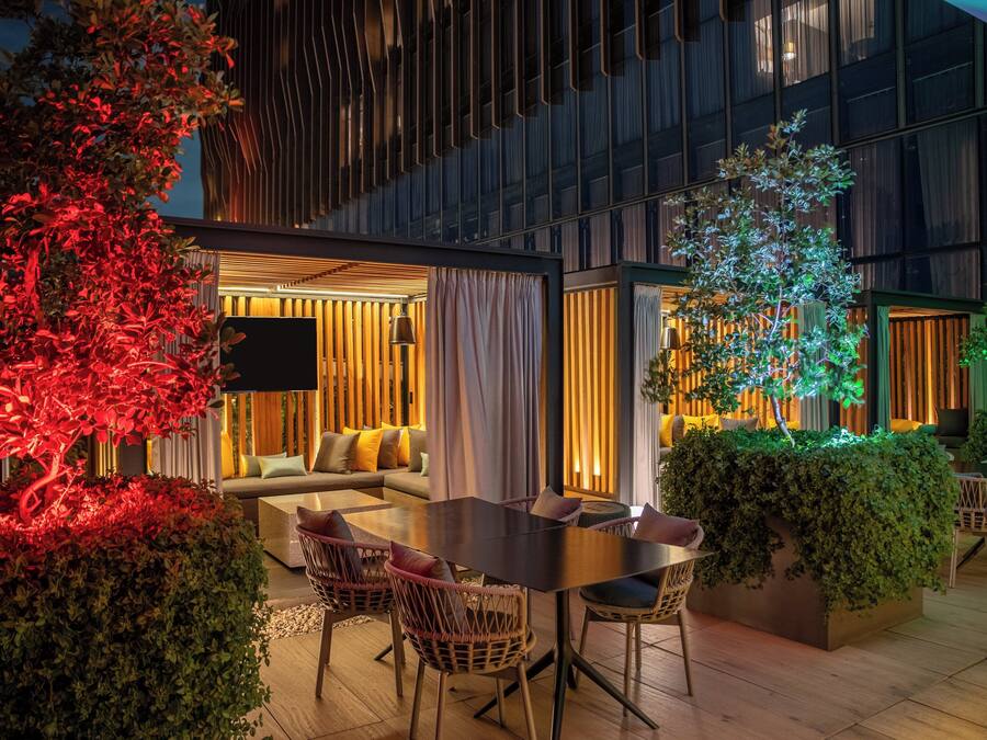 An outdoor dining area with a table and chairs, surrounded by greenery and a modern building in the background.