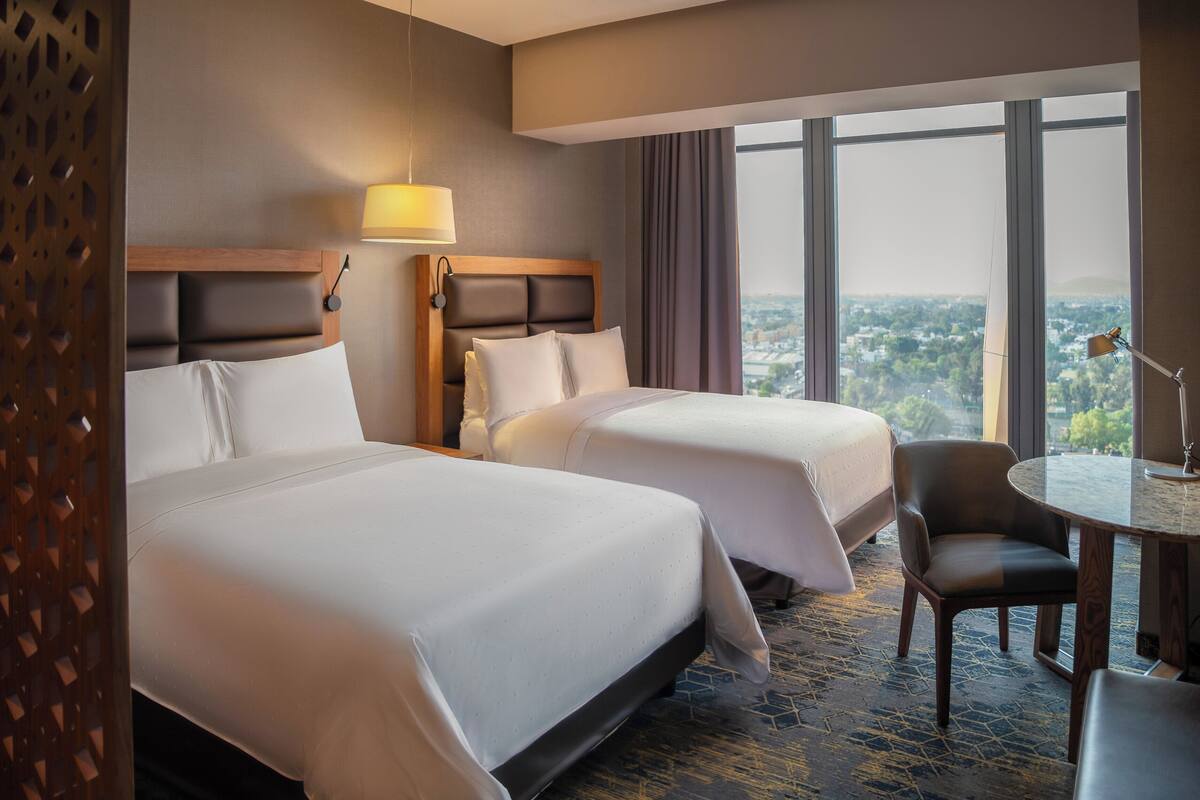 A hotel room with two beds, a desk, a chair, and a large window offering a city view.
