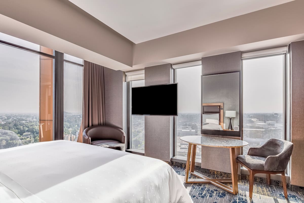 A hotel room with a large bed, a desk, a chair, a TV, and a view of the city.