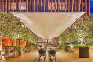 Un espace de restauration en plein air, aménagé avec des tables et des chaises, entouré de verdure et surmonté d’une grande structure en forme de tonnelle.