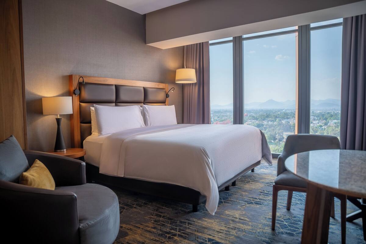 A hotel room with a large bed, a sitting area with a chair and a table, a lamp, and a view of a cityscape through large windows.