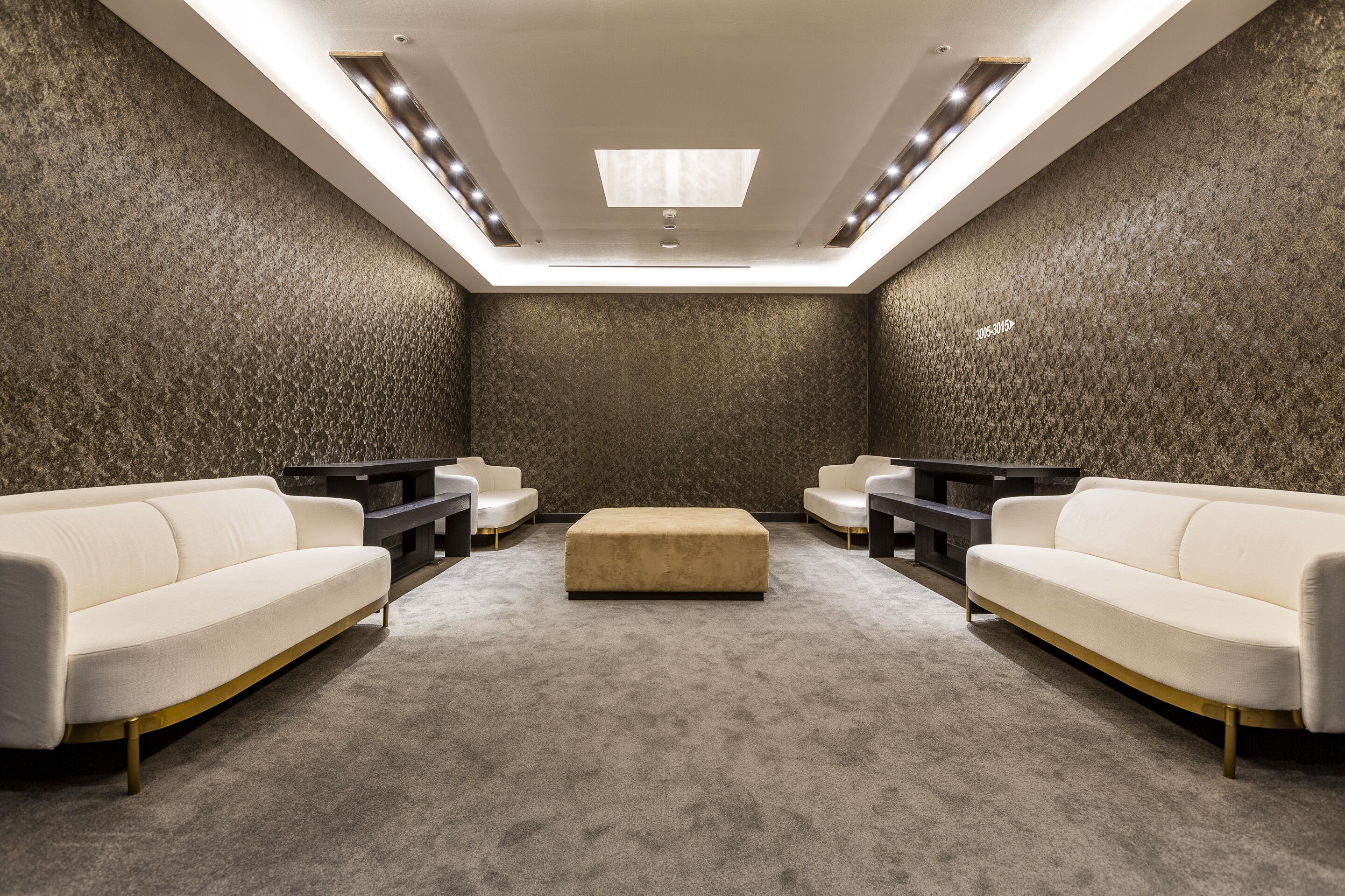 A modern, well-lit room with a high ceiling, a central ottoman, and two sofas.