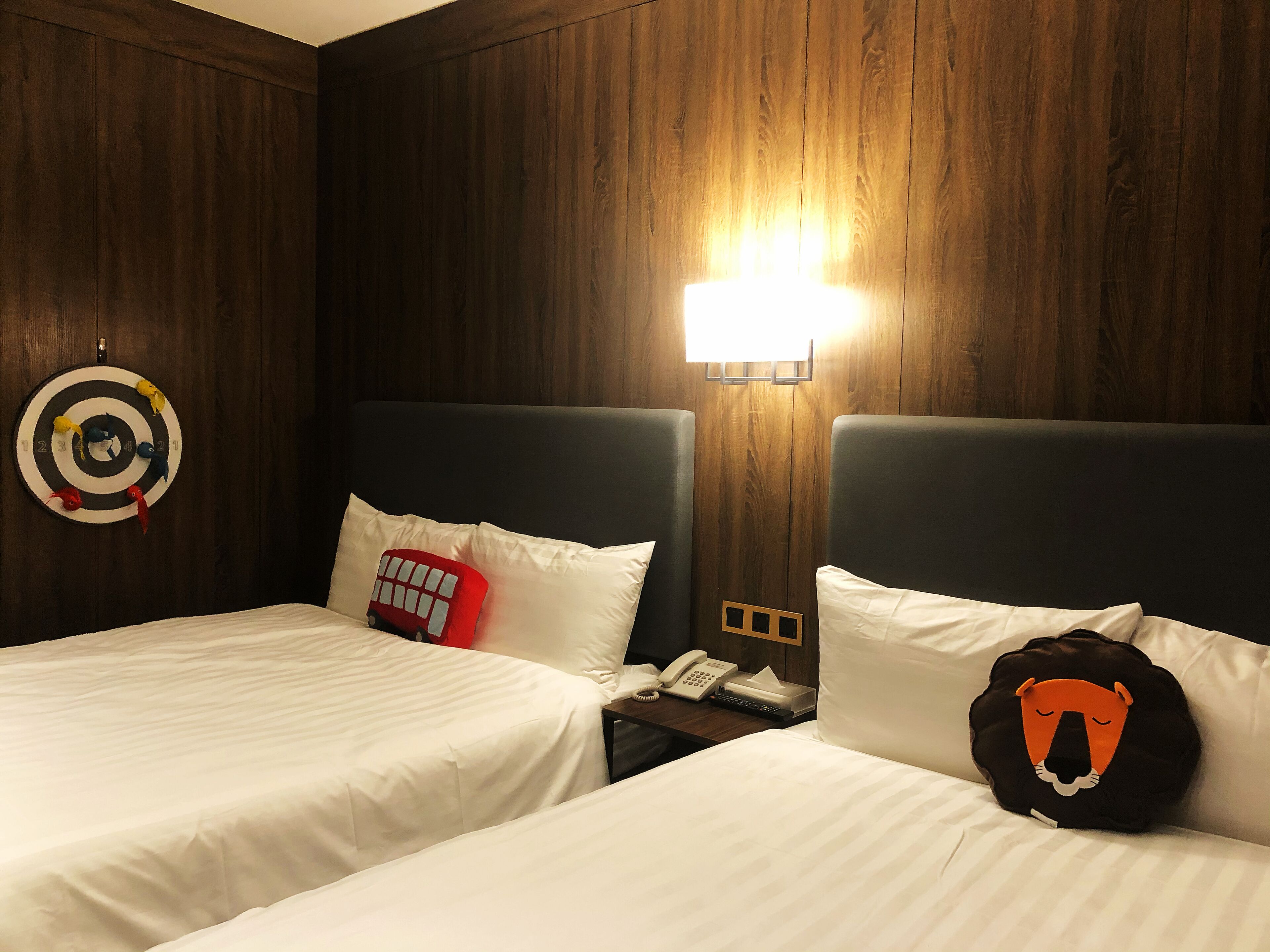 A hotel room with two beds, wooden paneling, a wall-mounted target, a lamp, and a lion-shaped pillow.