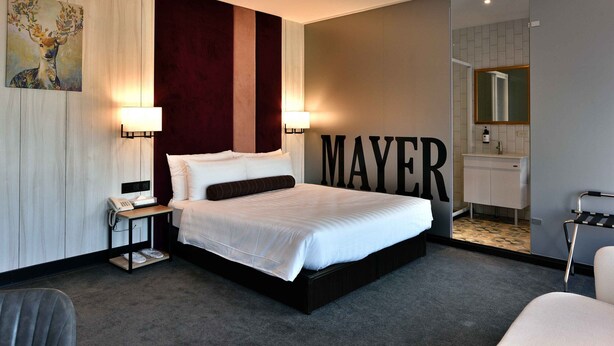 Une chambre d’hôtel moderne avec un grand lit, des lampes de chevet, un bureau avec un téléphone, un miroir et une salle de bain visible à travers une porte ouverte.