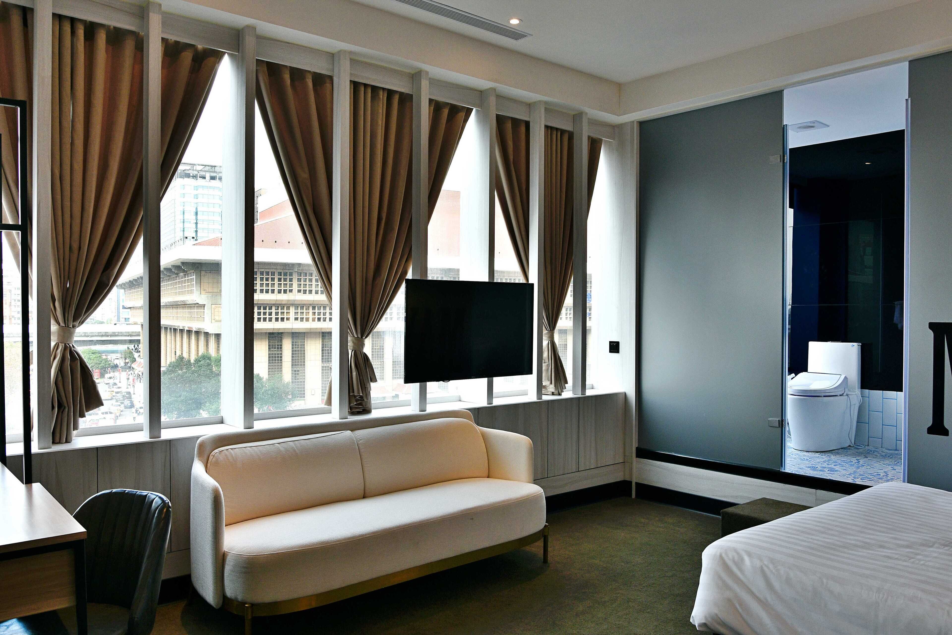 A hotel room with a large window, a sofa, a bed, a TV, and a view of the cityscape.