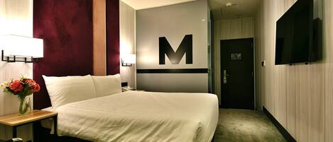 A hotel room with a large bed, a desk with a lamp, a vase of flowers, and a television on the wall.
