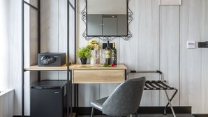 Une chambre d’hôtel compacte, avec un bureau en bois, un réfrigérateur noir, une chaise grise, une petite plante en pot, une bouteille de vin et un tableau encadré accroché au mur.