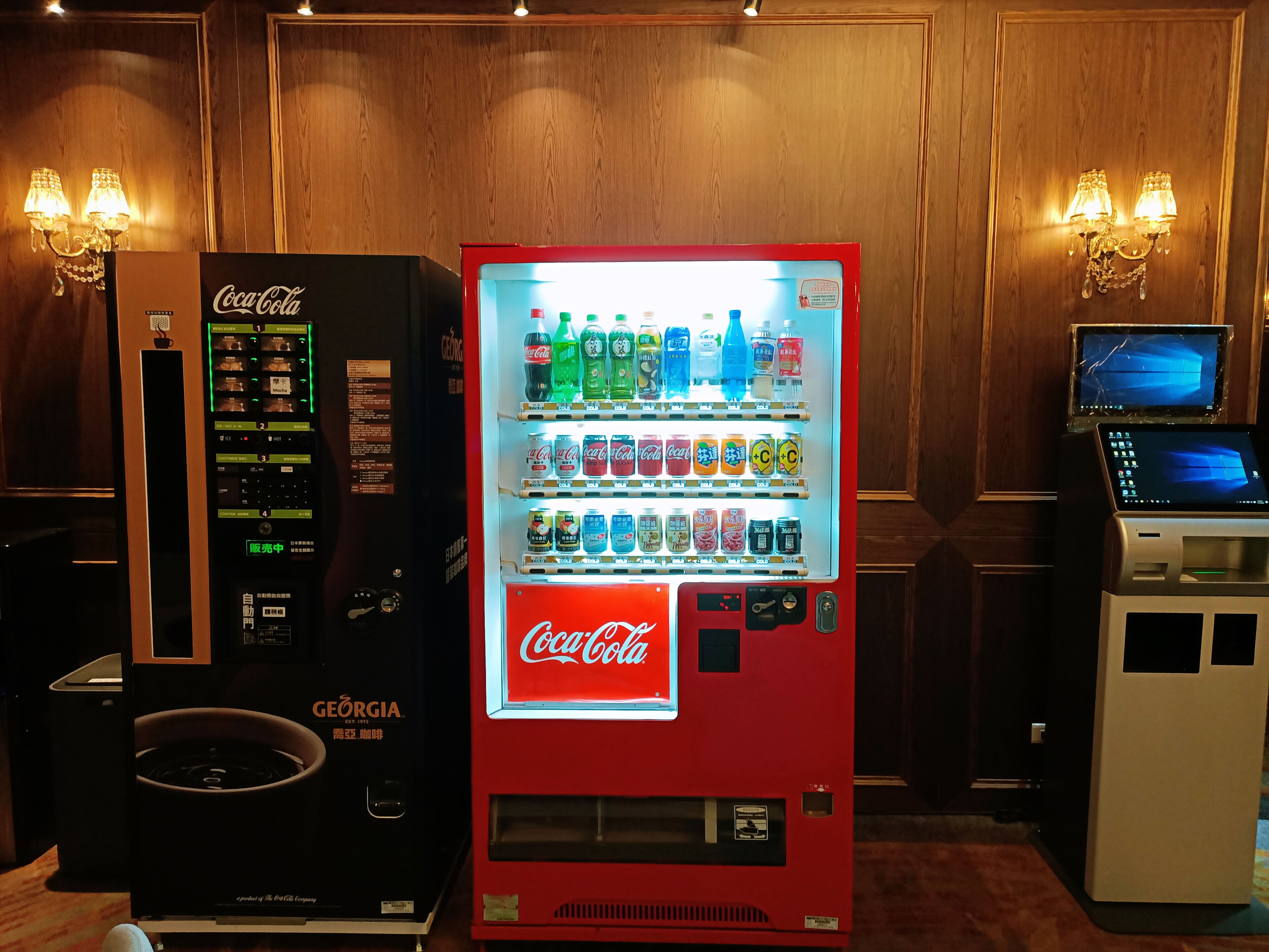 En Coca-Cola automtomat med et digitalt display, en kaffemaskine og en bærbar computer på et bord.