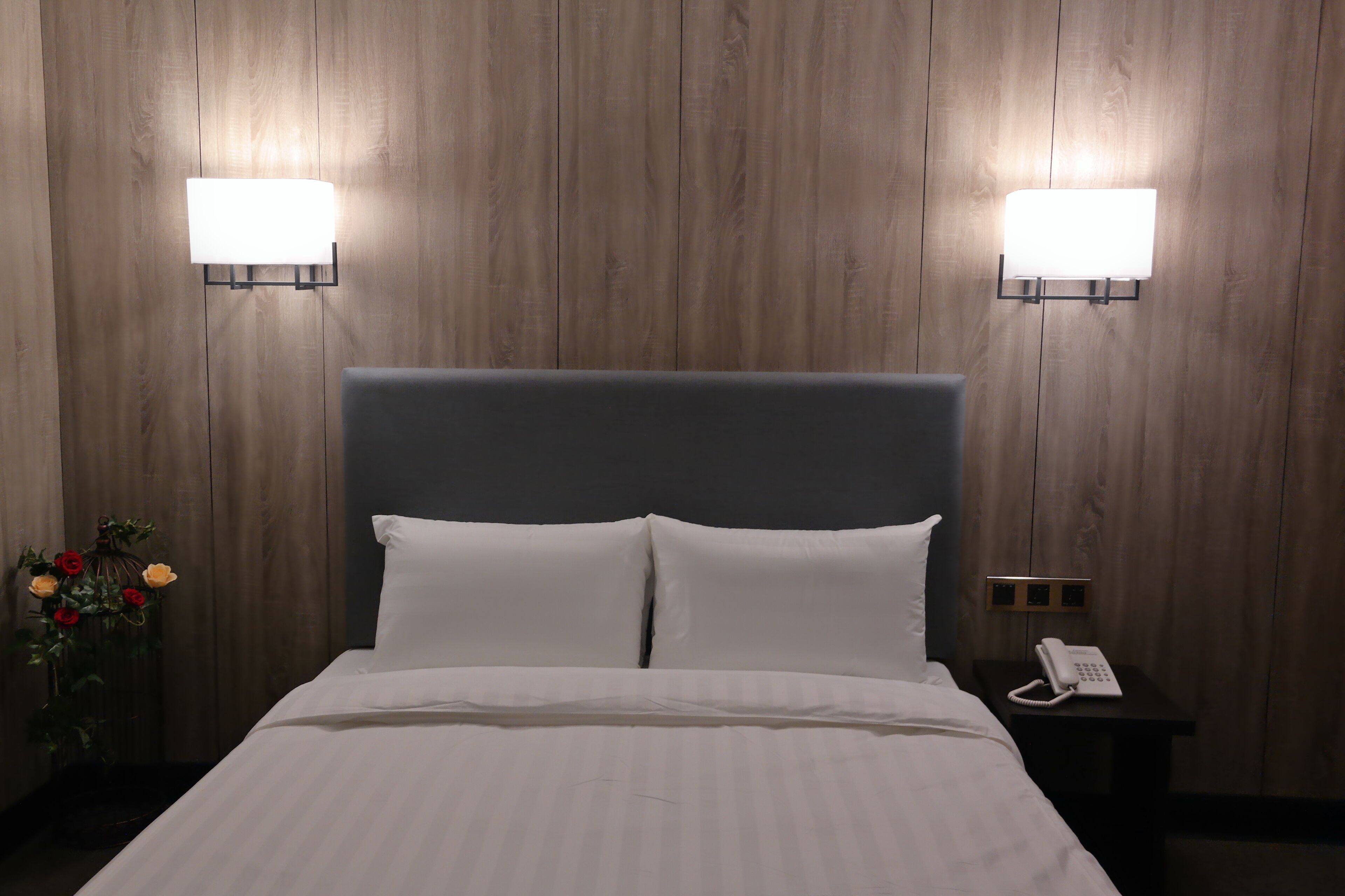 Ein Hotelzimmer mit einer Wandpaneele aus Holz, einem großen Bett mit weißen Leinen, zwei Wandleuchten, einem Nachttisch mit weißem Telefon und einer Vase mit Blumen.