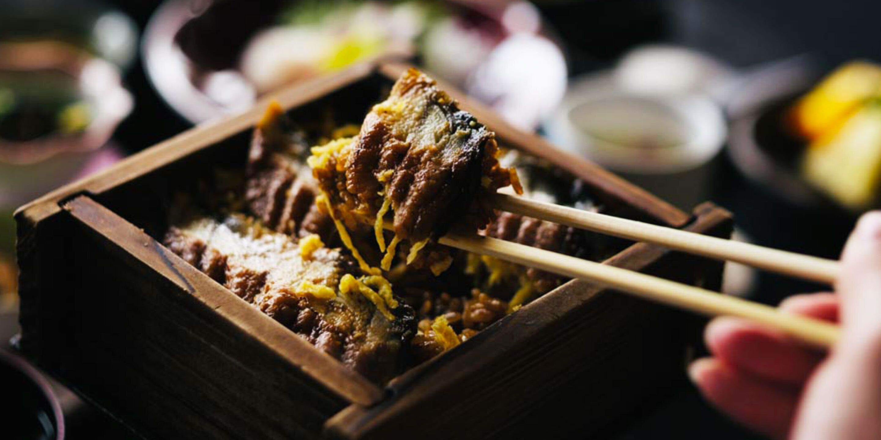 A close-up of a dish being held with chopsticks, possibly a type of meat dish, with a wooden tray in the background.