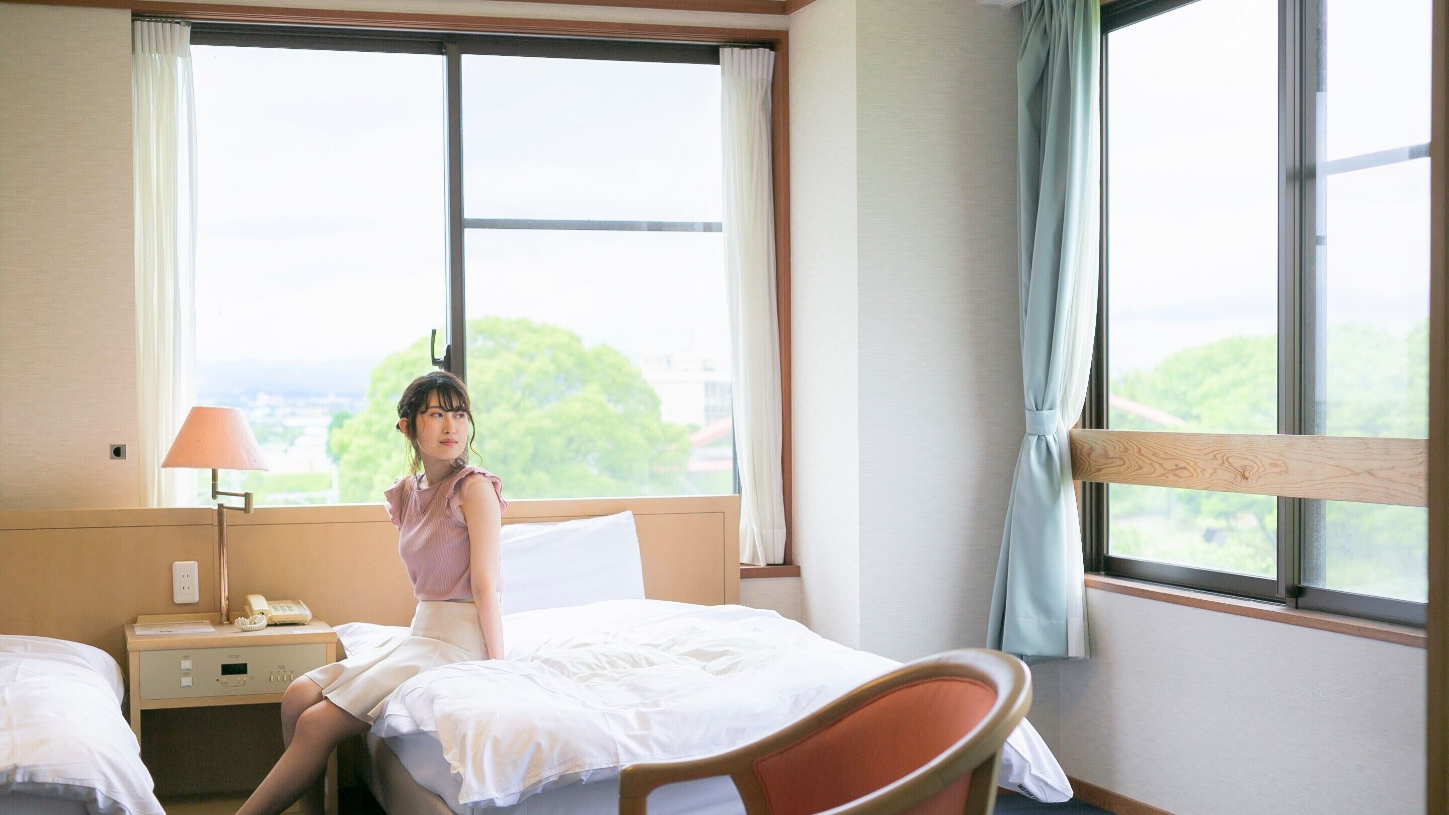 Een hotelkamer met een bed, nachtkastje met een lamp, een stoel en grote ramen met gordijnen.