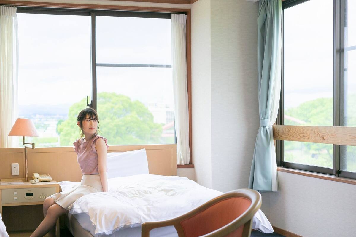 Une chambre d’hôtel avec un lit, une table de chevet avec une lampe, une chaise et de grandes fenêtres avec des rideaux.