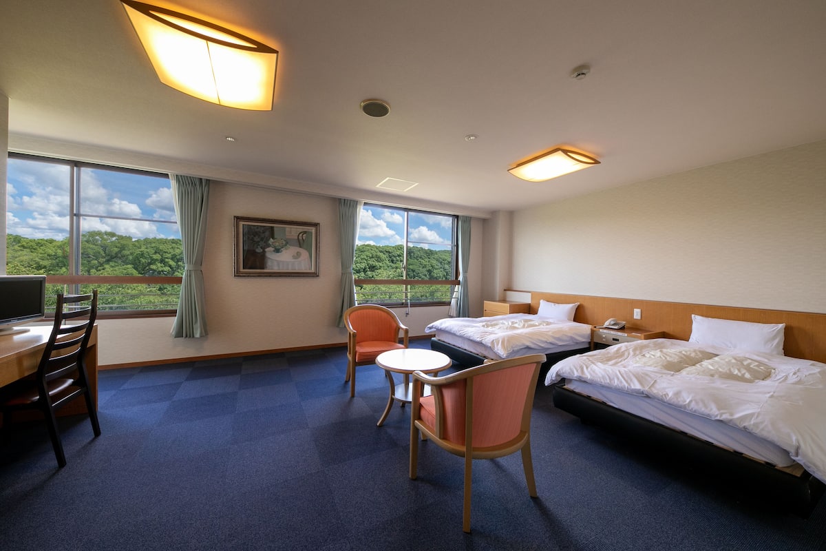 Une chambre d’hôtel avec deux lits, un bureau, une chaise et de grandes fenêtres donnant sur un paysage verdoyant.